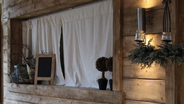 A rustic wooden cabin interior with a dining table set for two, a small window with curtains, and a wall-mounted light fixture. - B&B Ca' Virò (Albosaggia)