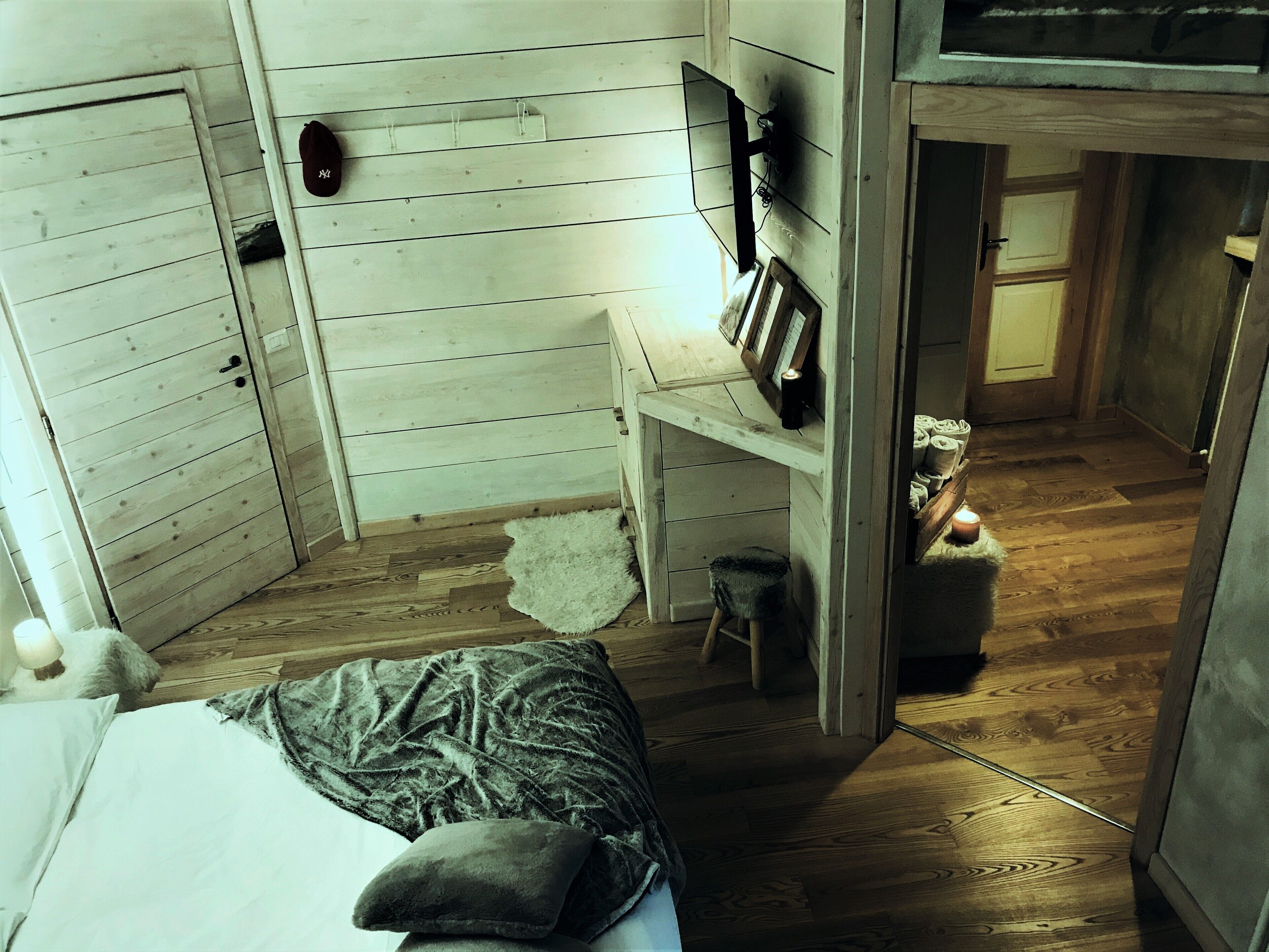 Une pièce chaleureuse aux murs en bois, avec un lit recouvert d’oreillers, un petit tabouret, une étagère ornée de cadres et une bougie allumée.