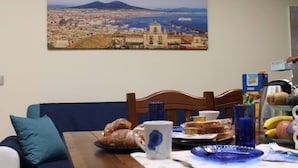 A dining area with a table set for breakfast, a cityscape painting on the wall, and a view of a coastal city.