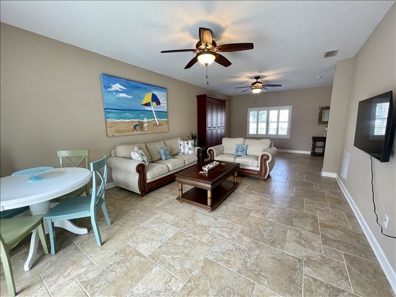 A spacious living room with a beige sofa, a coffee table, and a dining area with a table and chairs. There are two ceiling fans, a painting of a beach scene, and a TV mounted on the wall.