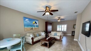 A spacious living room with a beige sofa, a coffee table, and a dining area with a table and chairs. There are two ceiling fans, a painting of a beach scene, and a TV mounted on the wall.