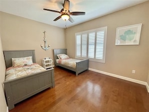 Quarto com duas camas, um ventilador de teto, uma janela com persianas e um adorno fixado na parede.
