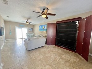 A spacious living room with a large sliding door, a ceiling fan, and a beach-themed painting on the wall. - Beach Bird House- Walk to the Beach! 4224 Oriole (Wilbur by the Sea)