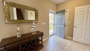 A hallway with a wooden console table, a decorative sailboat, a mirror with a geometric frame, and a door leading outside.