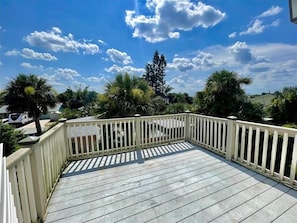 A wooden deck with a white railing, surrounded by palm trees and a clear blue sky with clouds.