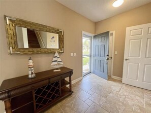 A hallway with a wooden console table, a decorative sailboat, a mirror with a geometric frame, and a door leading outside.
