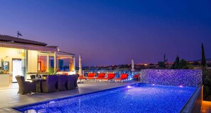 A poolside area with lounge chairs and a bar, overlooking a cityscape at dusk. - Stunning 3 bedroom villa 'JZ02' with private pool, beautiful interiors, communal pool and resort facilities, Zephyros Village, Aphrodite Hills (Kouklia)