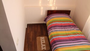 A small, narrow room with a wooden floor, a bed with a colorful striped bedspread, and a 'RELAX' mat in front of the bed.