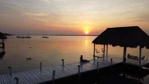 Zonsondergang vanaf een houten steiger met een rieten dakgebouw, boten en stilstaand water.