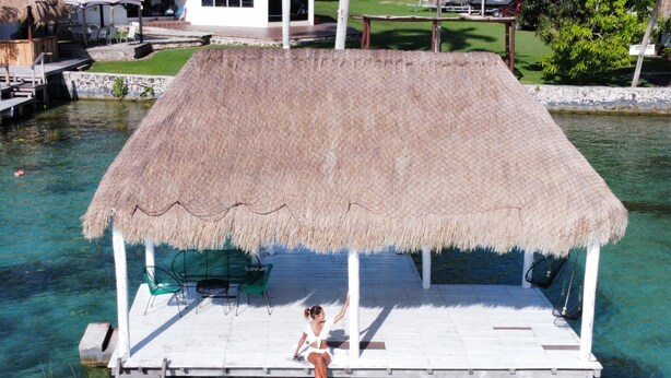 Een rieten dak bungalow op palen met een zwembad, lounge stoelen en een klein terras.