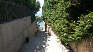 A narrow pathway flanked by a concrete wall on one side and a stone wall with dense foliage on the other, leading towards a body of water.