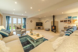 A cozy living room with a sofa, armchair, and a coffee table. There is a fireplace, a television, and a window with curtains. - Field Corner, Docking, Norfolk (Station Road)