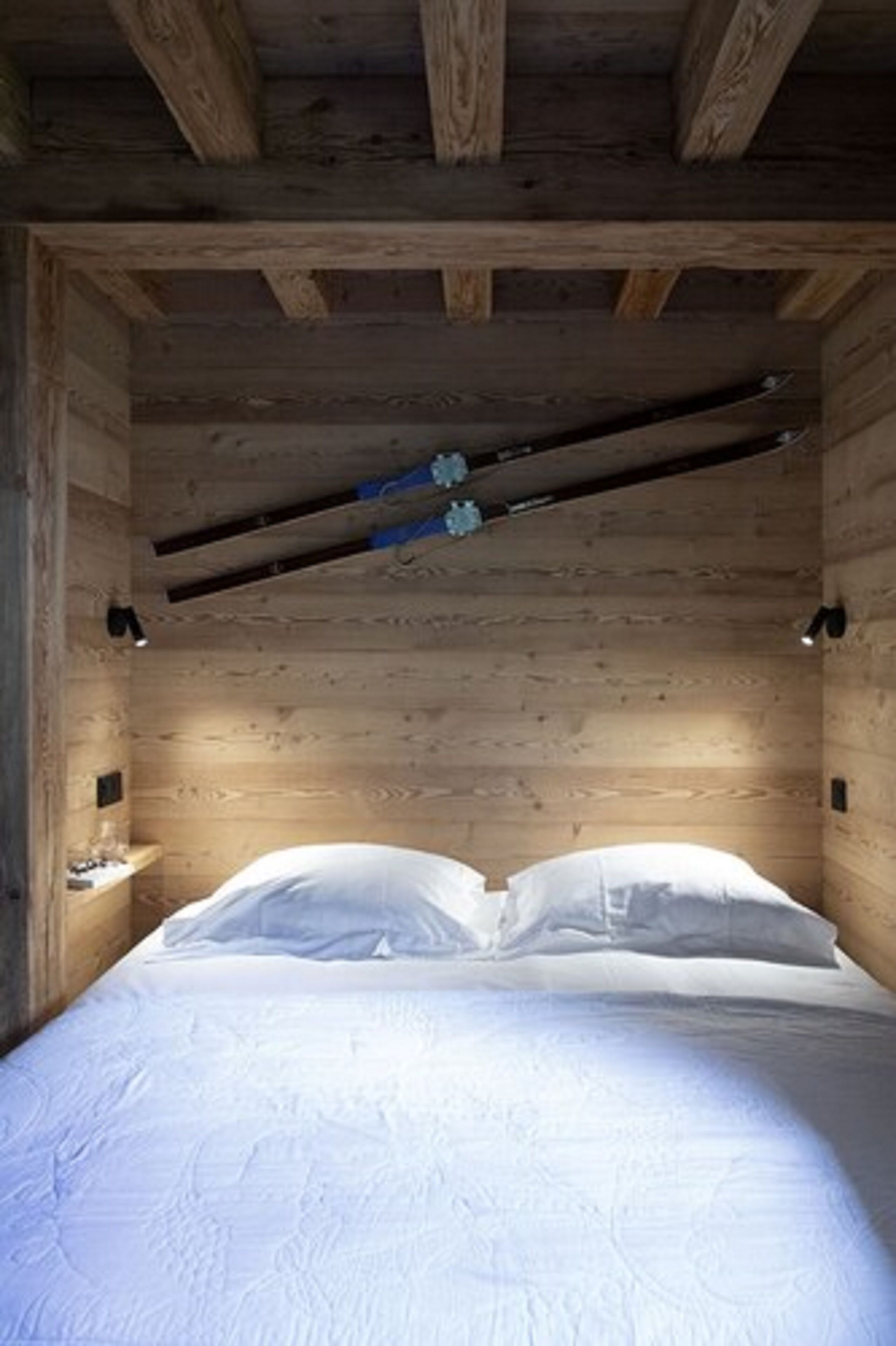 A wooden cabin-style bedroom with a bed centered under exposed beams, a small bedside table, and a wall-mounted light fixture.