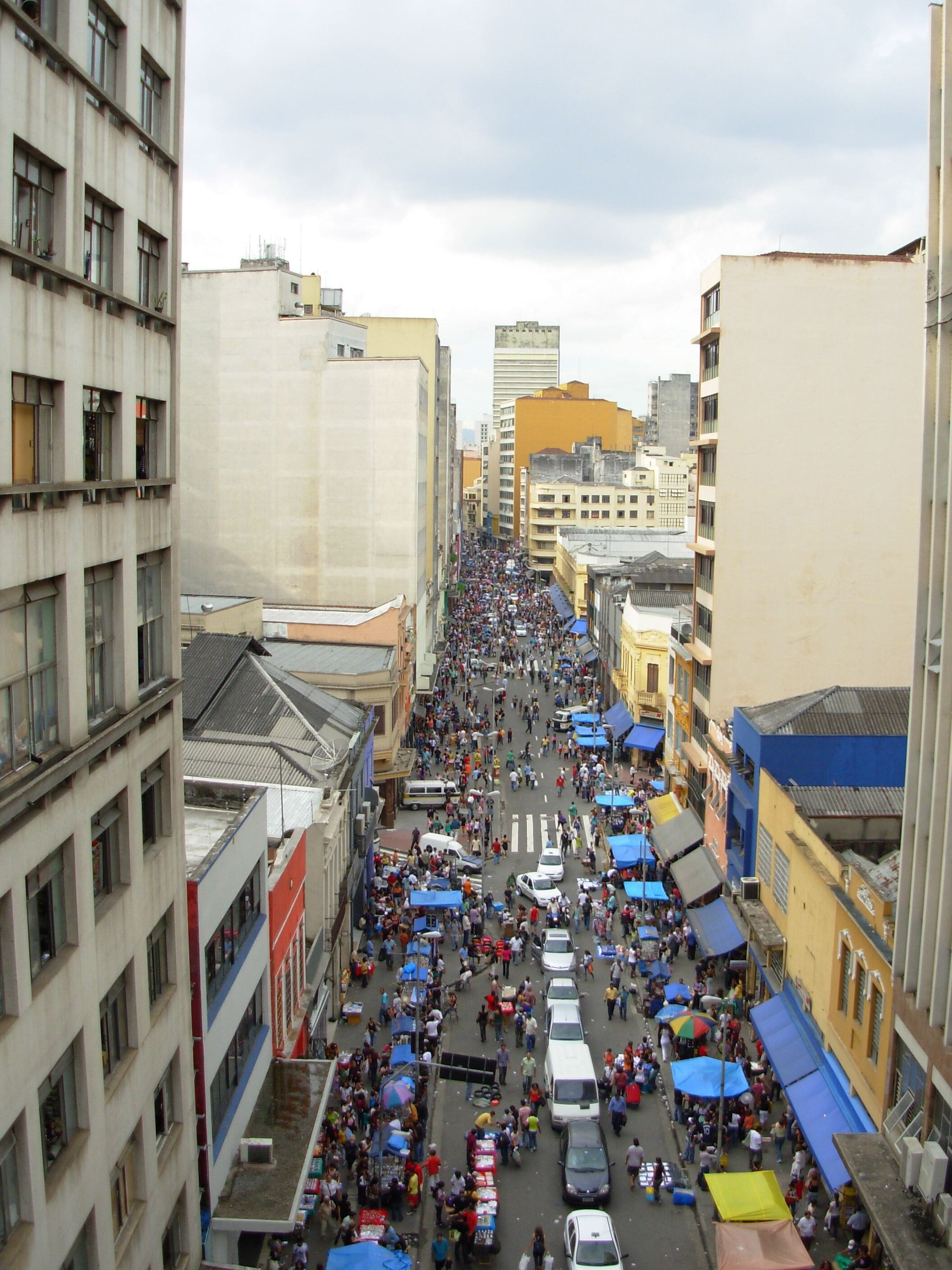 A crowded street with buildings on either side, a market with stalls, and a tall building in the background.