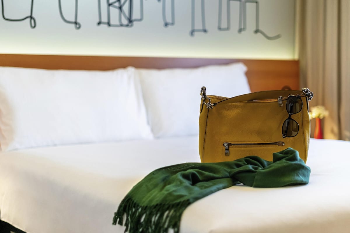 Una cama con ropa de cama blanca, un bolso amarillo y una manta verde.