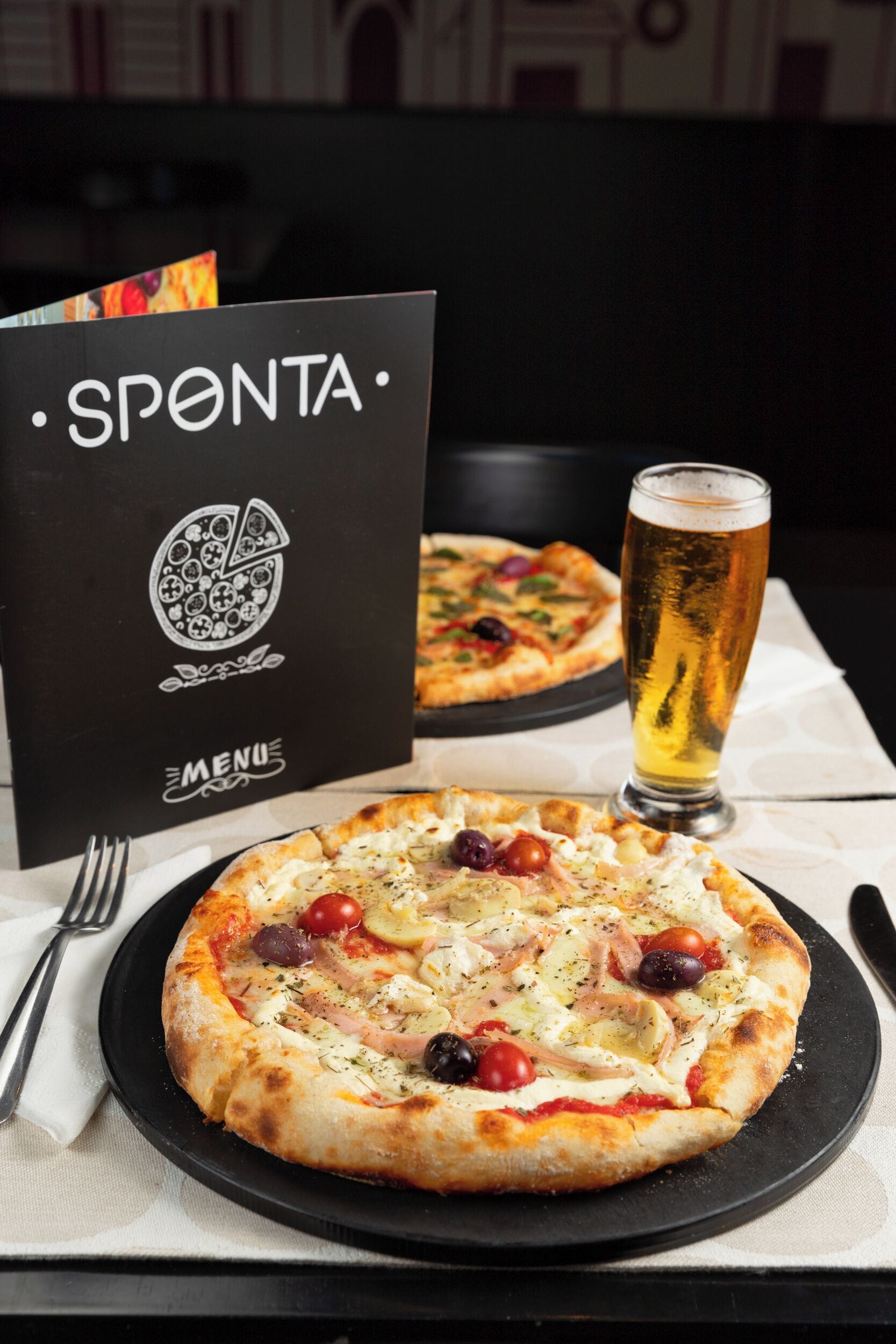 Pizza op een zwart bord met een menukaart en een glas bier op een tafel.