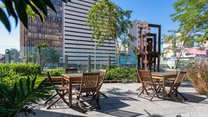 Een terras met houten tafels en stoelen, omringd door groen en moderne gebouwen op de achtergrond.