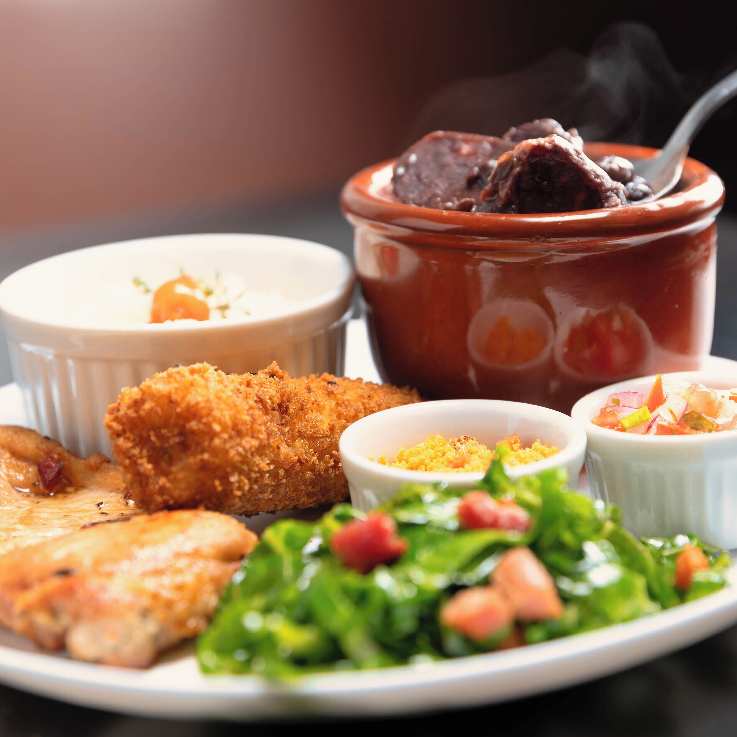 A plate with a variety of dishes including a breaded item, a bowl of soup, a salad, and two small bowls of condiments.