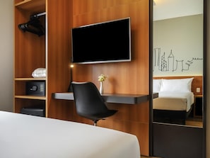 A hotel room with a bed, a TV mounted on the wall, a desk with a chair, a small safe, and a mirror reflecting the bed and a wall art piece. - Ibis Styles SP Centro (São Paulo)