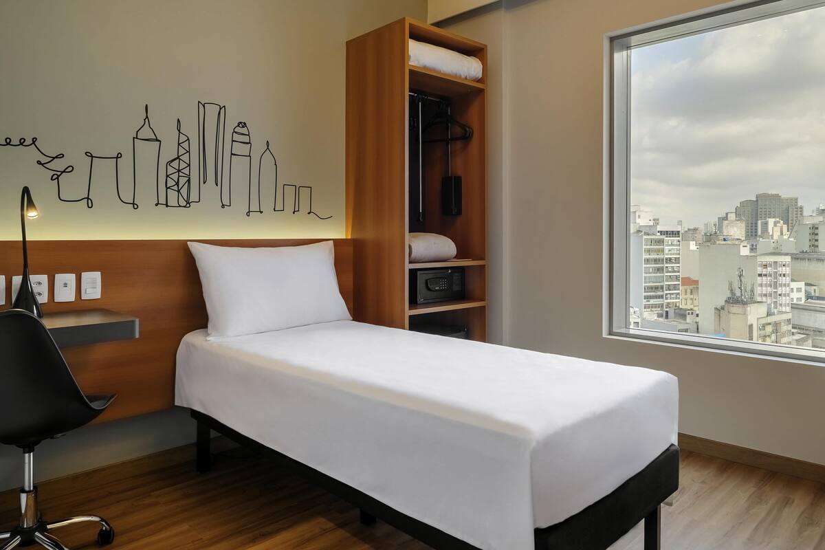 Una habitación de hotel con cama, escritorio, silla, armario y un gran ventanal con vistas a la ciudad.