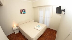 Ein Schlafzimmer mit Bett, Nachttisch, einem an der Wand montierten Fernseher und einem Fenster mit Vorhängen.