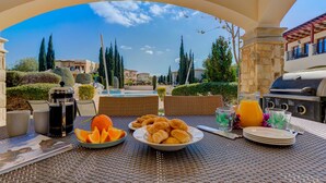 Un área de comedor al aire libre con una mesa dispuesta para el desayuno, incluyendo croissants y gajos de naranja, una parrilla y vistas de los edificios y el paisaje circundante.