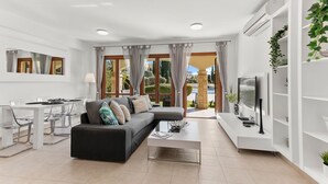 Una sala de estar moderna con un sofá seccional, una mesa de comedor con sillas, un televisor sobre un mueble blanco y un amplio ventanal con cortinas.