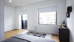 Un dormitorio moderno con un ventanal grande, una cama con colcha de estampado geométrico, una mesita de noche pequeña con objetos decorativos y un armario empotrado.