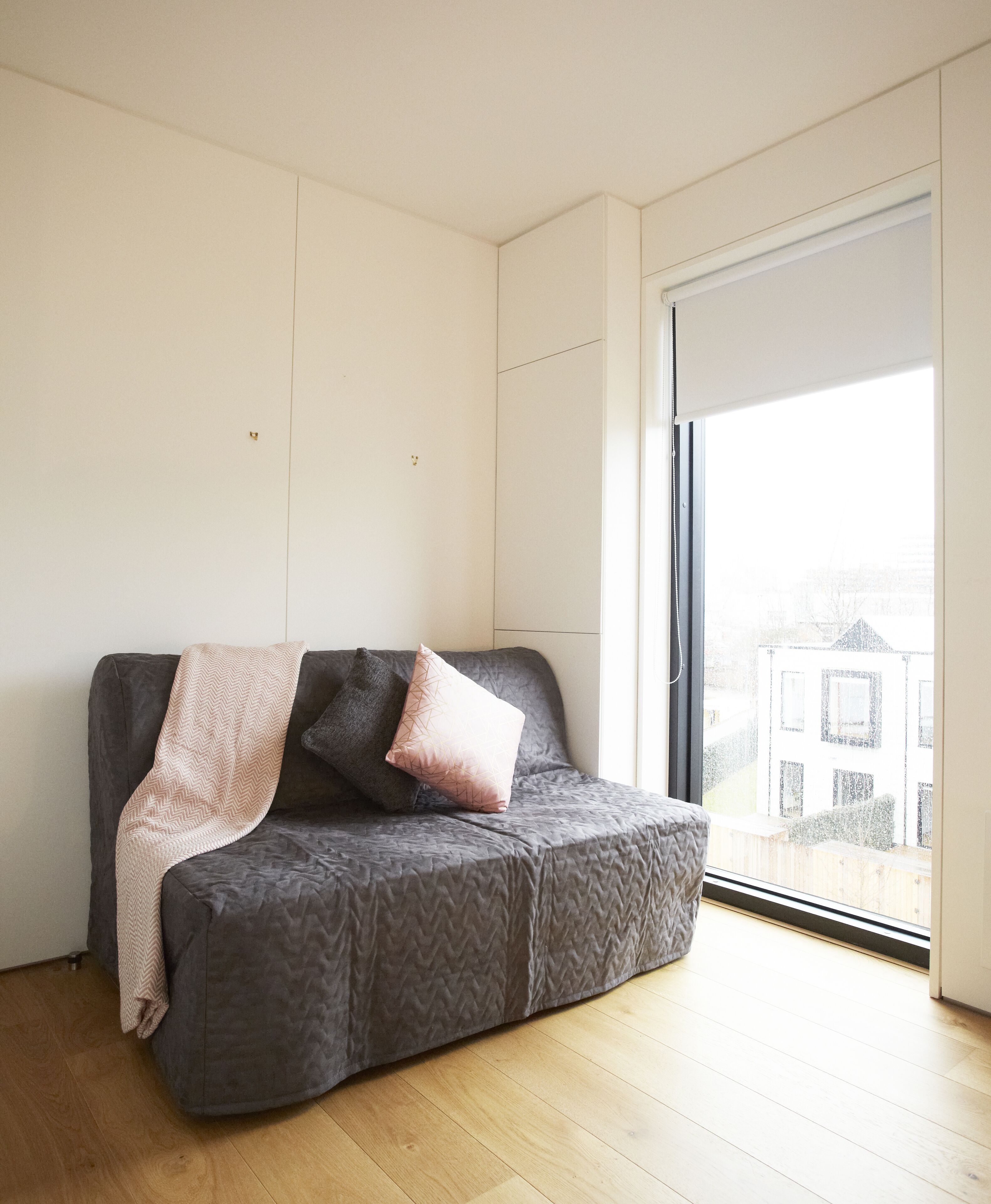 A modern living room with a grey sofa, a pink throw blanket, and a large window offering a view of buildings.
