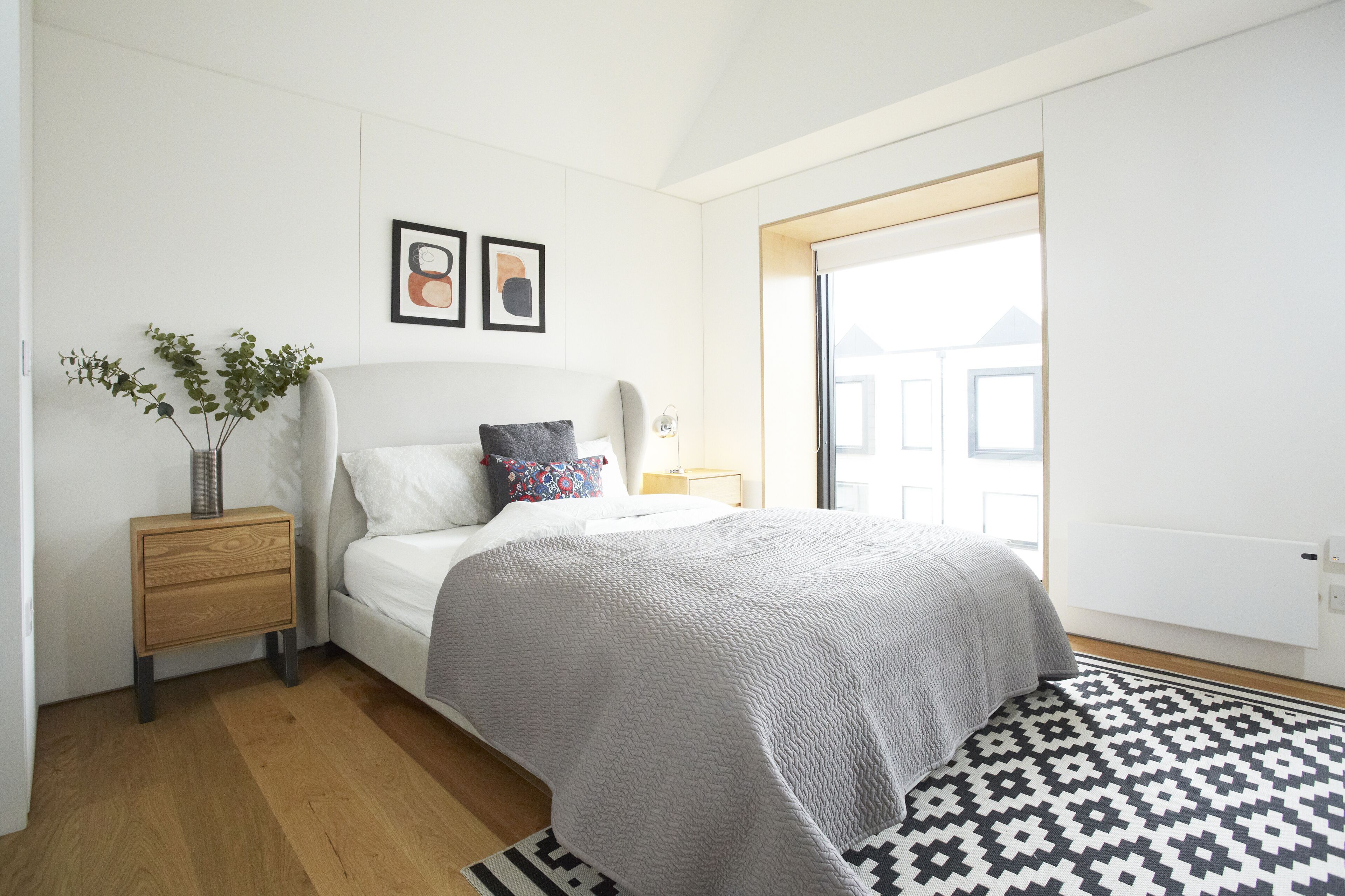 A modern bedroom with a large bed, a bedside table, a nightstand, a floor lamp, and a window with a view of buildings.