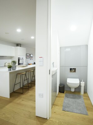 Een moderne keuken met witte kasten, een barstoel en een kleine plant. Grenzend daaraan een badkamer met toilet, wastafel en een spiegel.