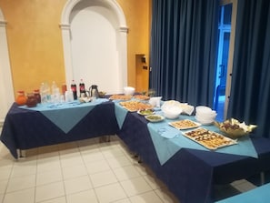 Une table de buffet garnie de divers plats et boissons, placée devant un mur jaune orné d’une porte en arc de cercle.