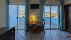 Une pièce comprenant une table à manger, deux chaises, un téléviseur fixé au mur et une porte-fenêtre coulissante donnant sur un balcon avec vue sur la ville.