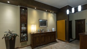 A reception area with a wooden reception desk, a potted plant, a framed picture, and a yellow radiator. - Azzurro Hotel Residence (L'Aquila)
