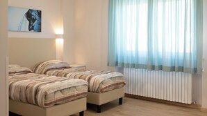 A bedroom with two beds, a wall-mounted artwork, a bedside lamp, and a window with sheer curtains. - Azzurro Hotel Residence (L'Aquila)
