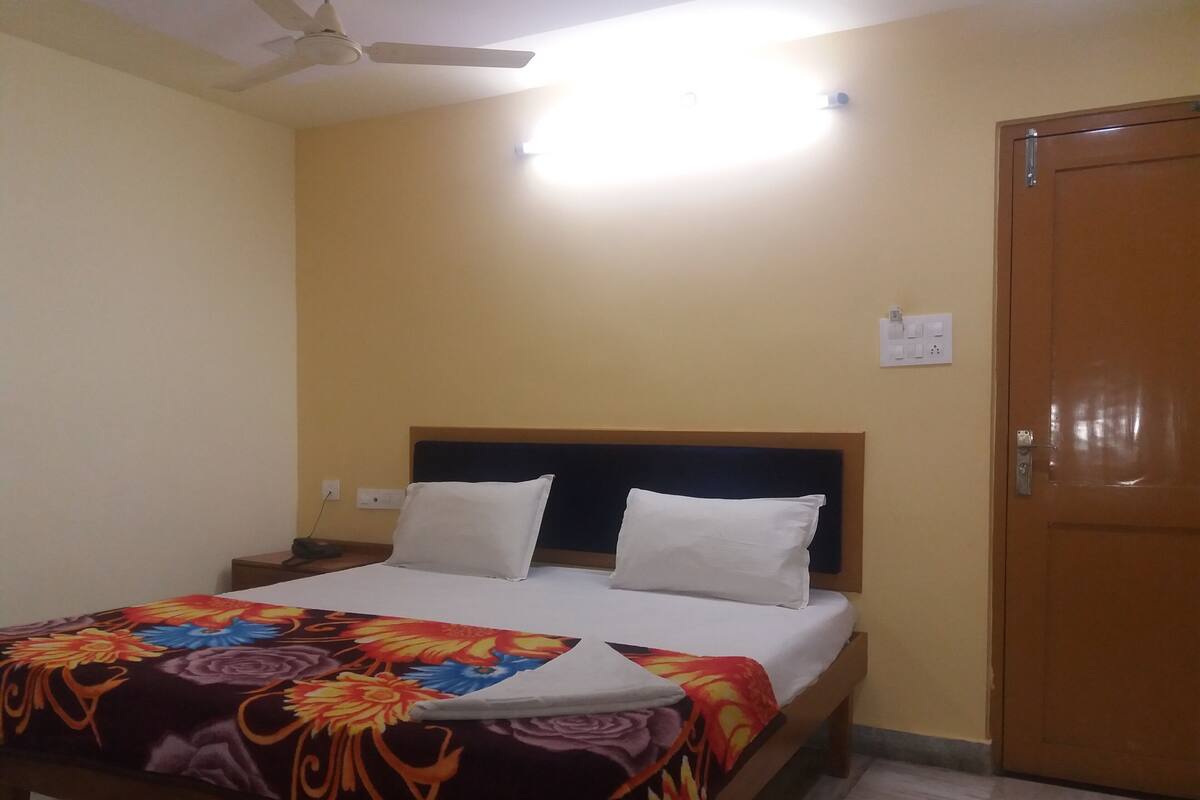 Una habitación de hotel con cama, ventilador de techo, puerta de madera y mesita de noche.