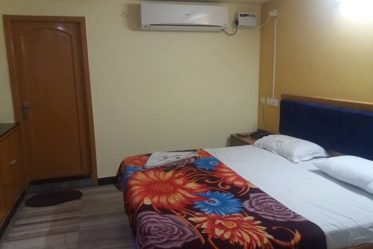 Una habitación de hotel con una cama, una mesita de noche, un equipo de aire acondicionado y una puerta de madera.