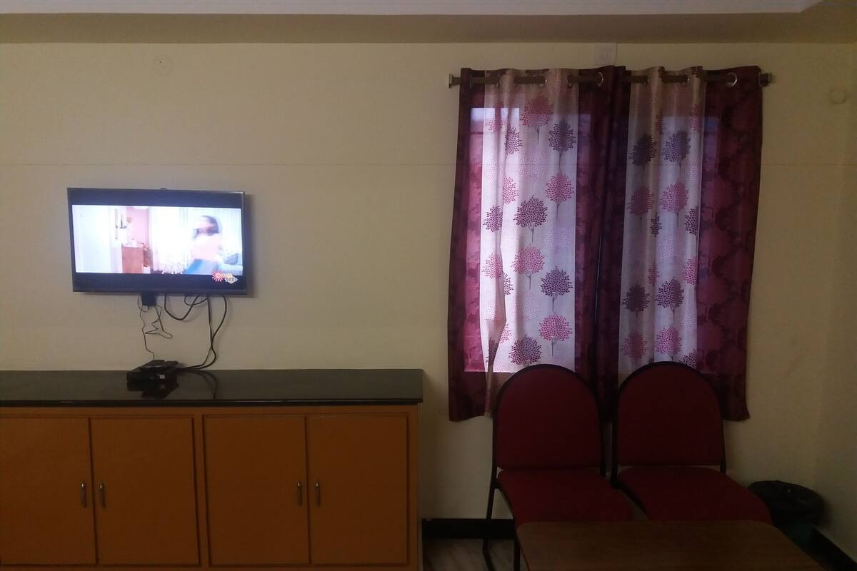 Una habitación con una televisión montada en la pared, un armario de madera, dos sillas rojas y una mesa.