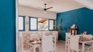 A dining area with white chairs and tables, a ceiling fan, and a view of the outside through large windows. - Pousada Costamar (Porto Seguro)