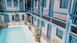 A pool area with a blue and white building, potted plants, and a tiled floor. - Pousada Costamar (Porto Seguro)