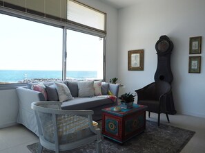 A modern living room with a large window overlooking the sea, a grey sofa with cushions, a wicker chair, a small table with a plant, and a grandfather clock.