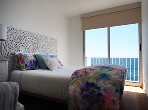 A bedroom with a bed, a nightstand, a lamp, and a view of the sea through a large window.