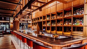 A bar area with wooden shelves stocked with bottles, a long counter with stools, and a visible kitchen area in the background. - Surety Hotel, Autograph Collection (Des Moines)
