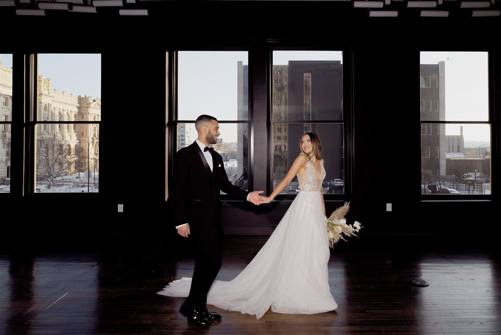 A couple holding hands in a room with large windows and a wooden floor.