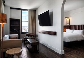 A hotel room with a bed, a sofa, a small table, a TV mounted on the wall, and a window with curtains.