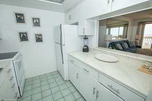 A compact kitchen with white cabinets, a refrigerator, a blender, and a dining area with a sofa and a large window.