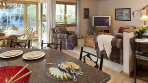 A cozy living room with a fireplace, a dining area with a table set for two, and a large window offering a view of the outdoors. - Shadowbrook (Snowmass Village)