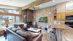 A spacious living room with a leather sofa, a stone wall, a fireplace, and a kitchen area with wooden cabinets and a microwave. - Shadowbrook (Snowmass Village)