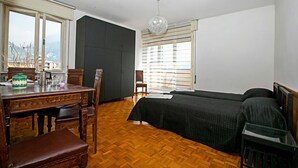 A bedroom with a wooden floor, a bed, a desk with a chair, a window with blinds, and a hanging light fixture.