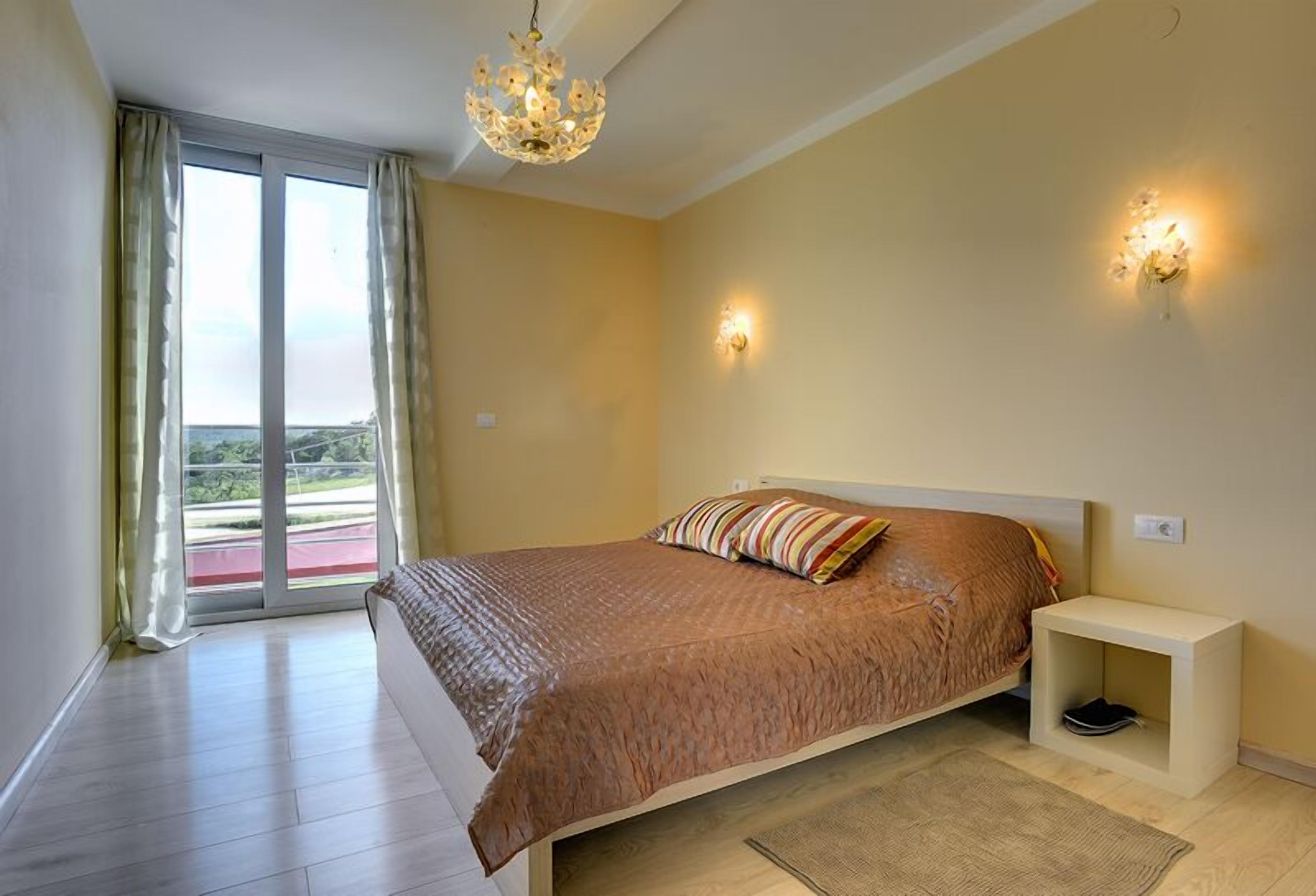 Une chambre à coucher avec un grand lit, une table de chevet, un lustre et une vue sur l’extérieur, accessible par une porte-fenêtre coulissante.
